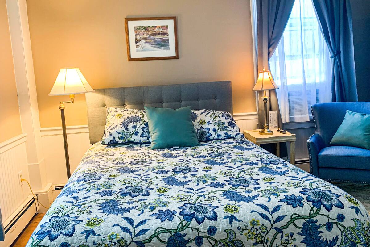 A bedroom with a bed, two bedside lamps, a chair, a framed picture on the wall, and a window with curtains.