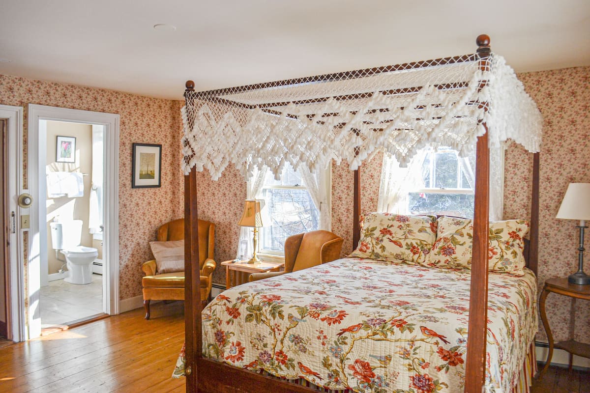 A bedroom with a four-poster bed, floral wallpaper, and a wooden floor.