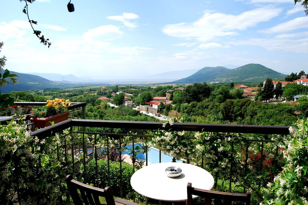 A balcony with a table and chairs overlooking a pool and a scenic view of a village and mountains.