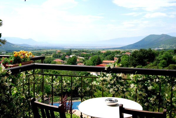 A balcony with a table and chairs, overlooking a scenic landscape with mountains and a village. - Jennifer Home Hotel (Drama)