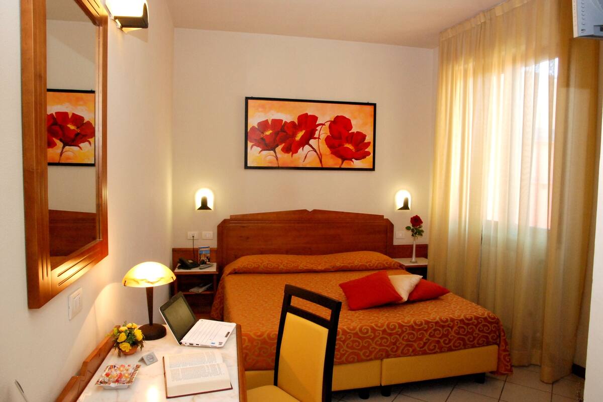 A hotel room with a bed, a desk with a laptop, a chair, a nightstand, a lamp, and a framed picture on the wall.