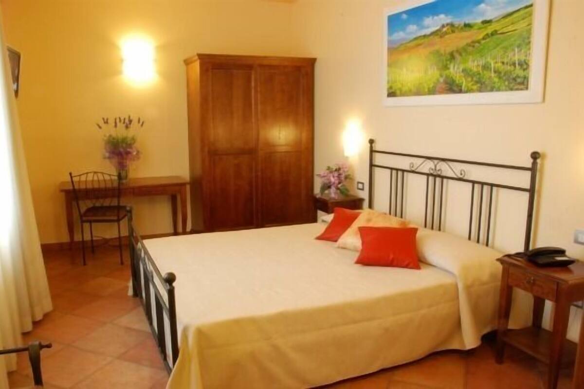 A bedroom with a bed, a wooden wardrobe, a desk with a chair, and a painting on the wall.