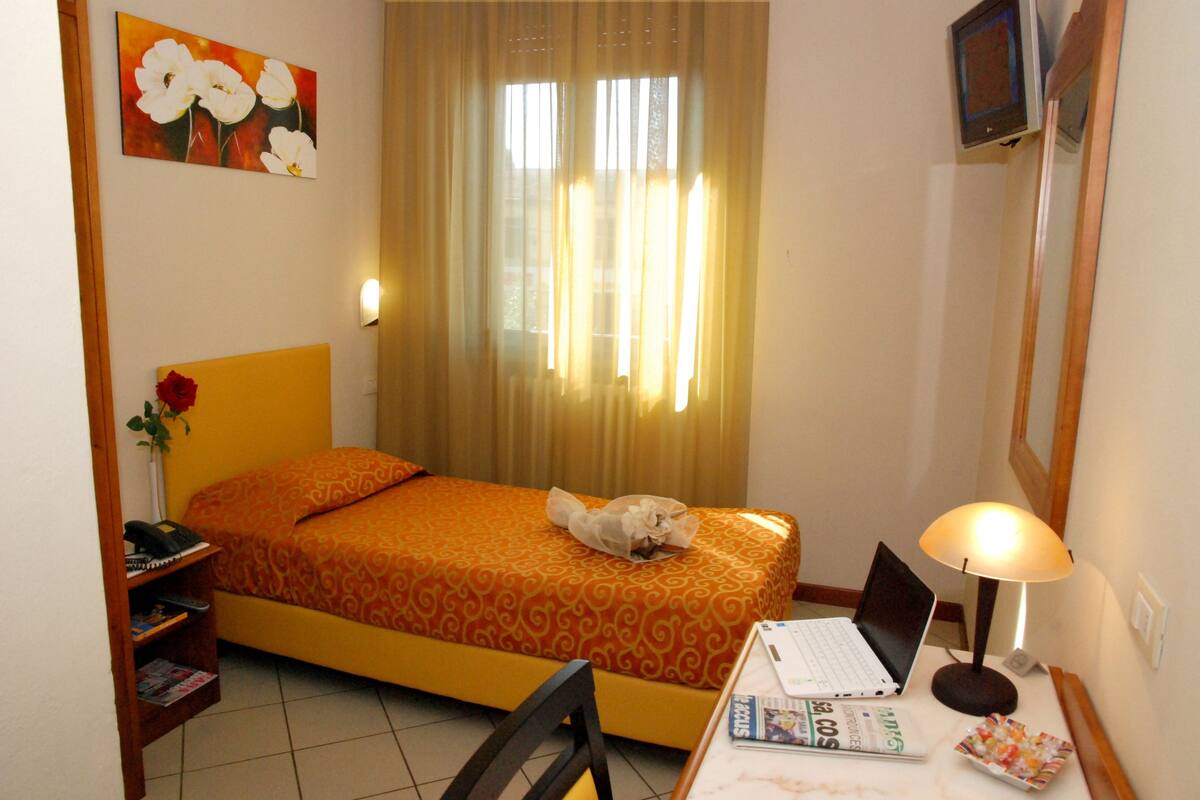 A hotel room with a bed, a desk with a laptop, a lamp, a chair, a phone, a vase with a flower, and a painting on the wall.