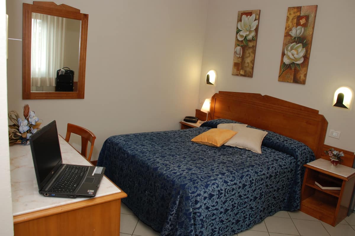 A bedroom with a bed, a desk with a laptop, a mirror, and two wall-mounted lamps.