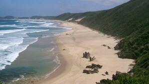 A coastal beach with a sandy shore, rocky outcrops, and green hills in the background.