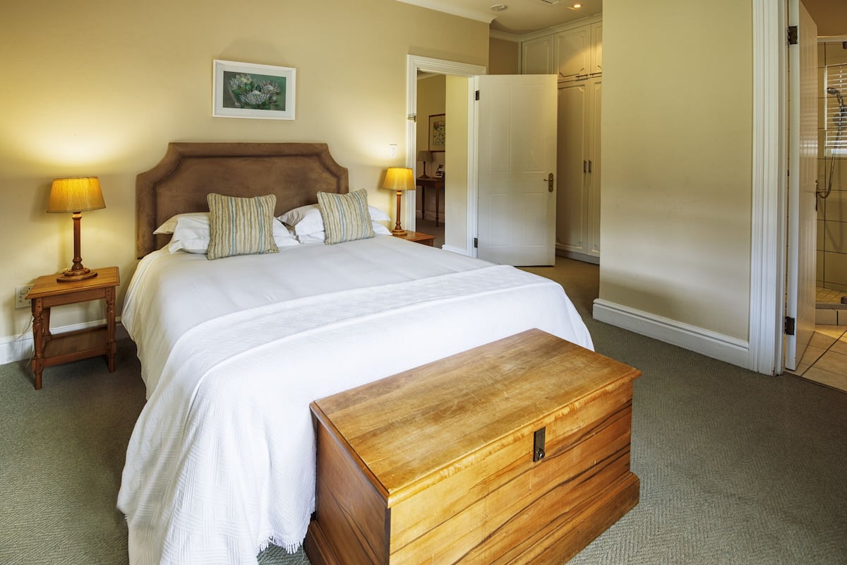 A neatly made bed with a wooden chest of drawers, a nightstand with a lamp, and a door leading to another room.
