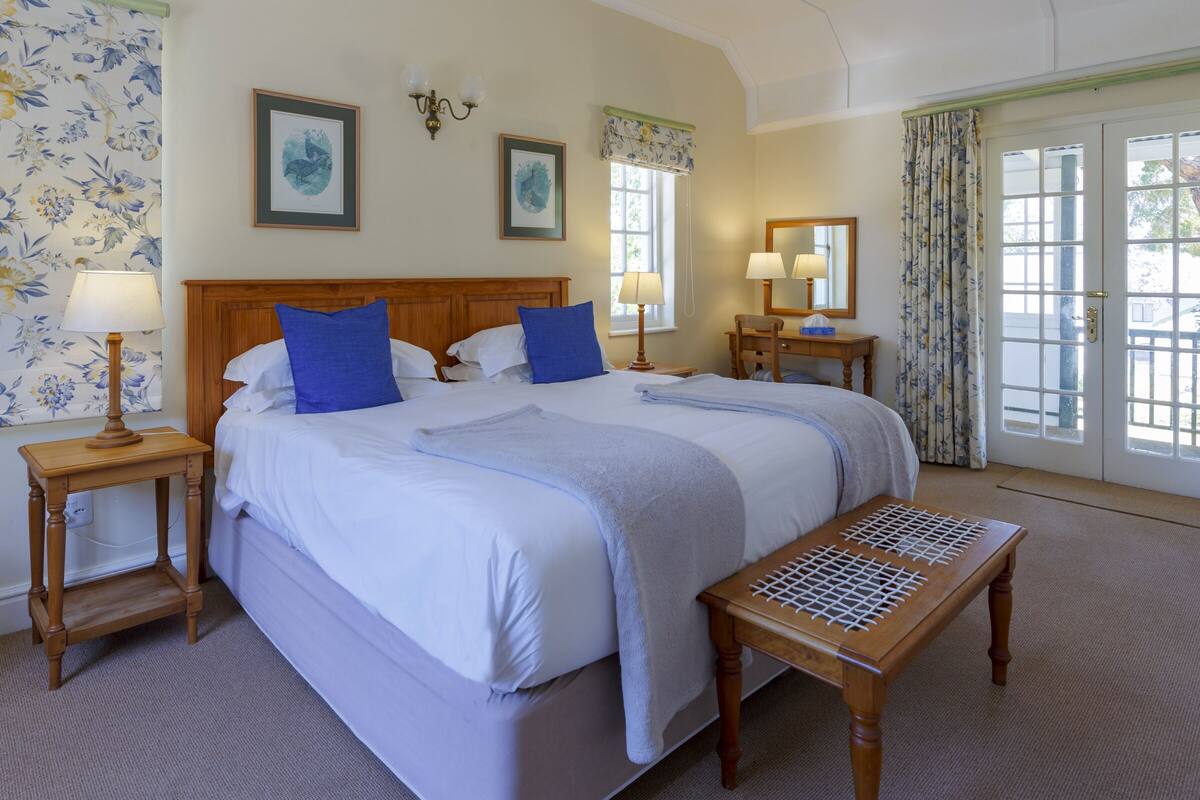 A double bed with a wooden headboard, two bedside tables with lamps, a wooden bench, and a large window with curtains.