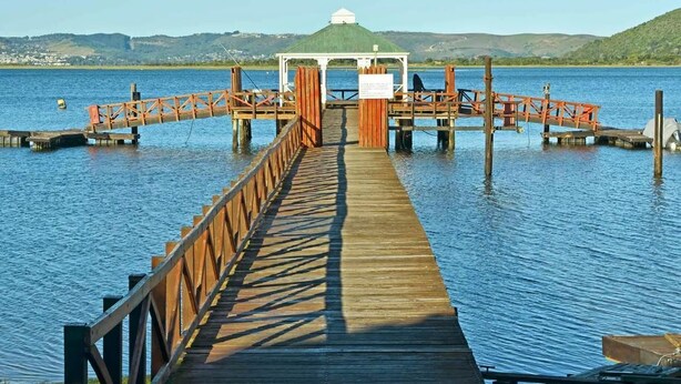 A wooden pier leading to a gazebo with a green roof, surrounded by calm water and distant hills.