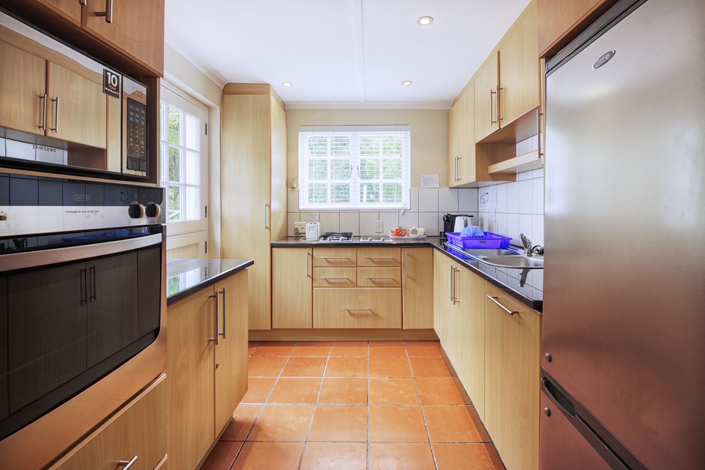 A modern kitchen with wooden cabinets, a stainless steel refrigerator, and a window with blinds.