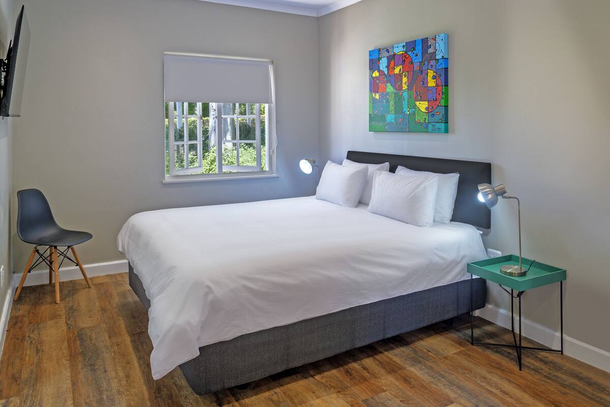 A bedroom with a bed, a chair, a bedside table with a lamp, a window, and a colorful abstract painting on the wall.