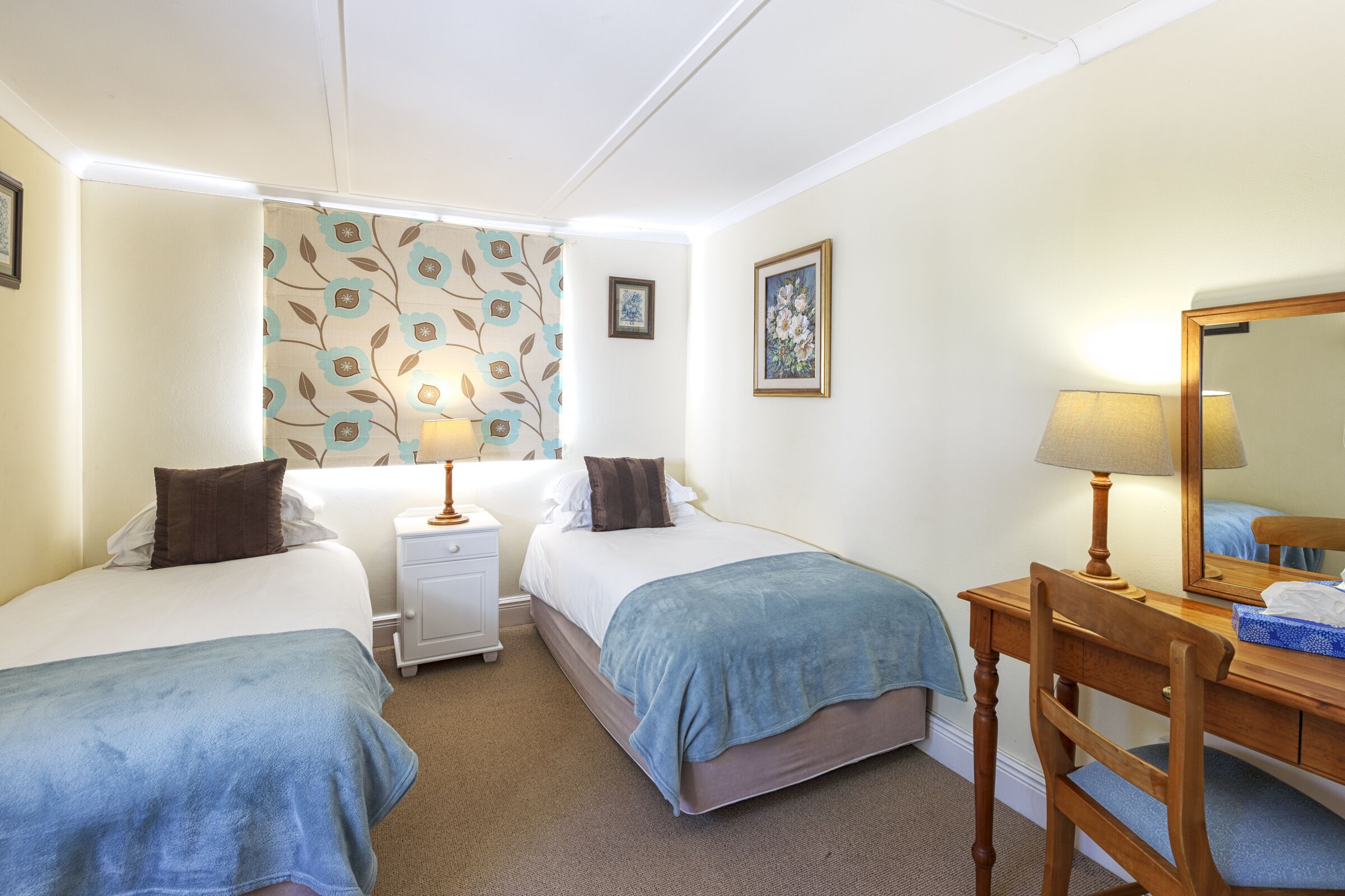 A bedroom with two beds, a desk with a chair, a mirror, and a nightstand with a lamp.