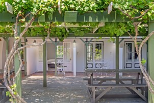 Un espacio de estar al aire libre cubierto con bancos y una mesa, rodeado por una pérgola con enredaderas.