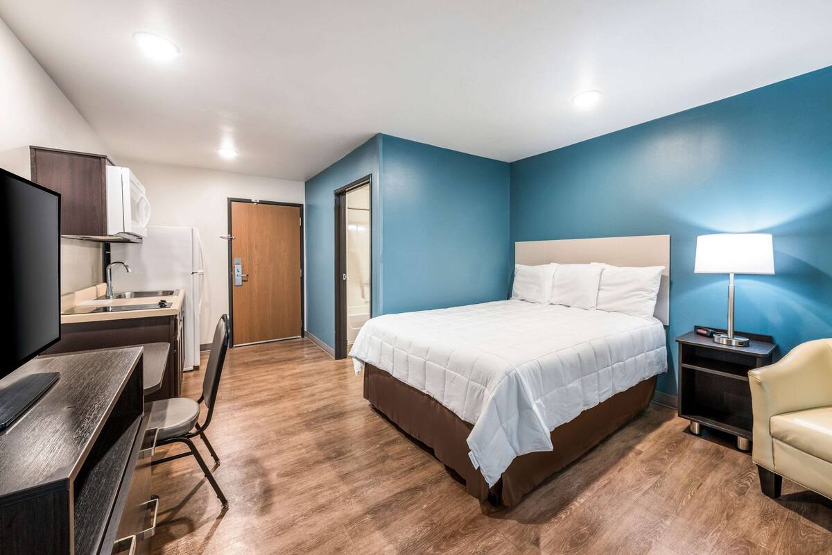 A hotel room with a bed, a desk, a chair, a TV, a lamp, and a bathroom door.