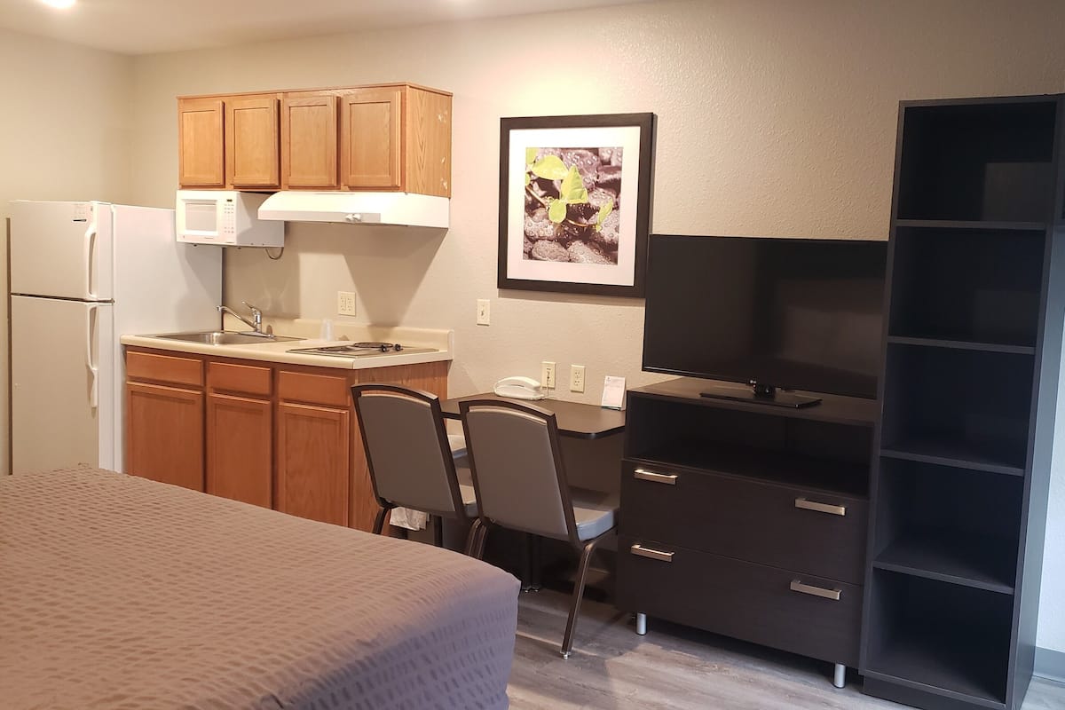 A compact kitchen with wooden cabinets, a microwave, and a refrigerator. There is a dining area with a table and chairs, and a black entertainment unit with a TV.