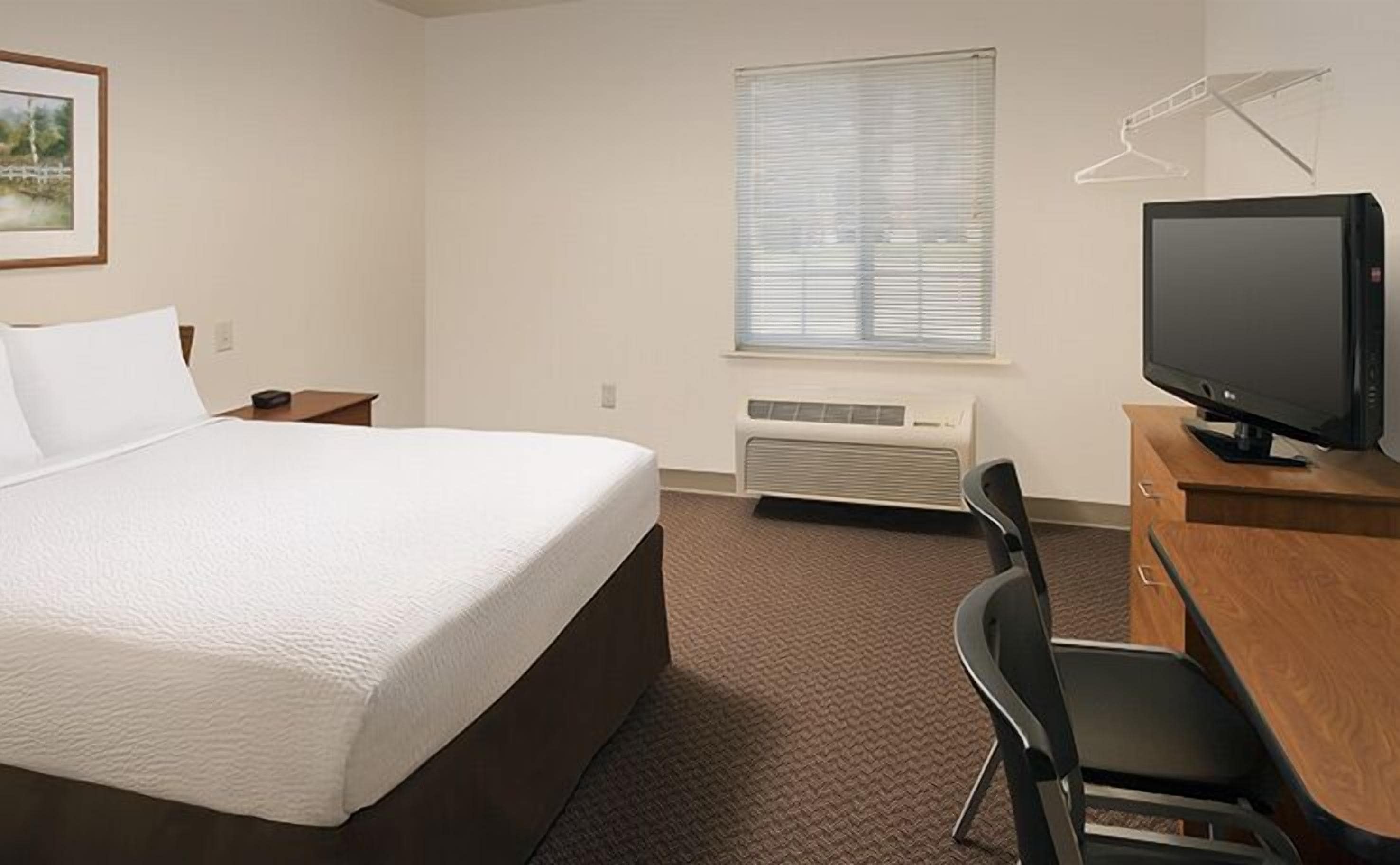 A hotel room with a bed, a desk, a chair, a television, a window with blinds, and a picture on the wall.