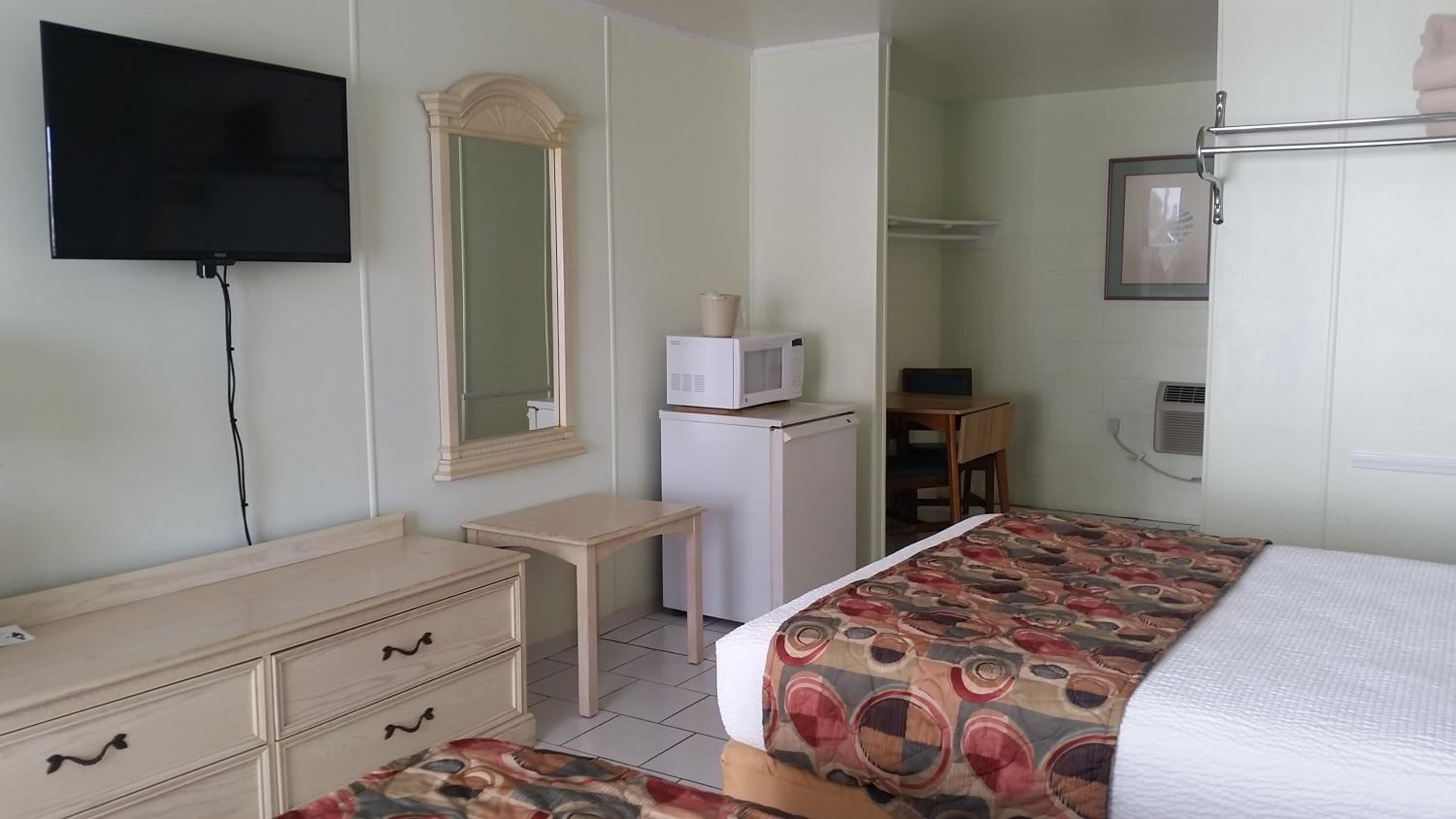 Une chambre d’hôtel comprenant un lit, une commode, un bureau, un miroir, une télévision, un four à micro-ondes et une armoire.