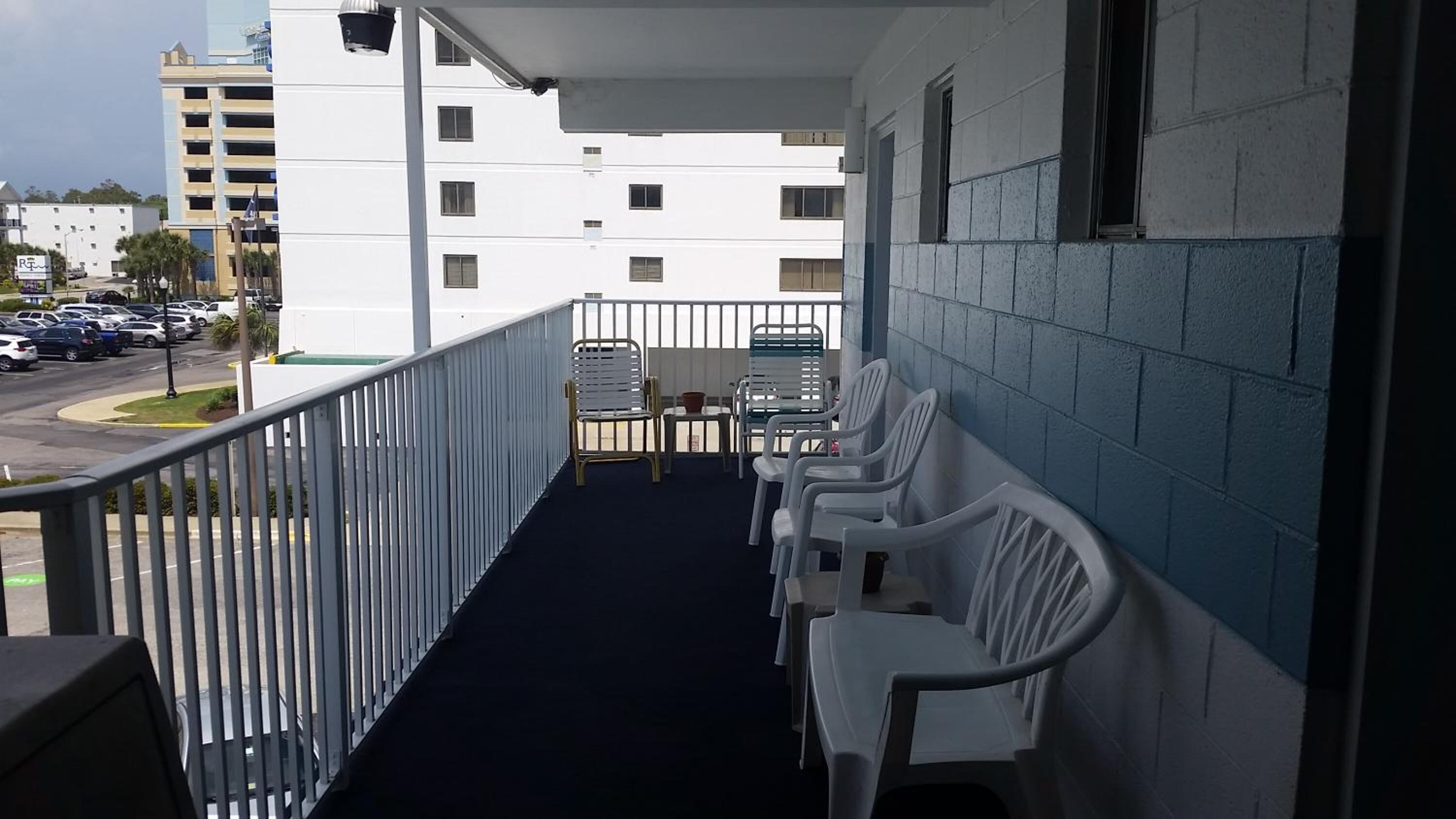 A balcony with white plastic chairs, a metal railing, and a view of a parking lot and buildings.