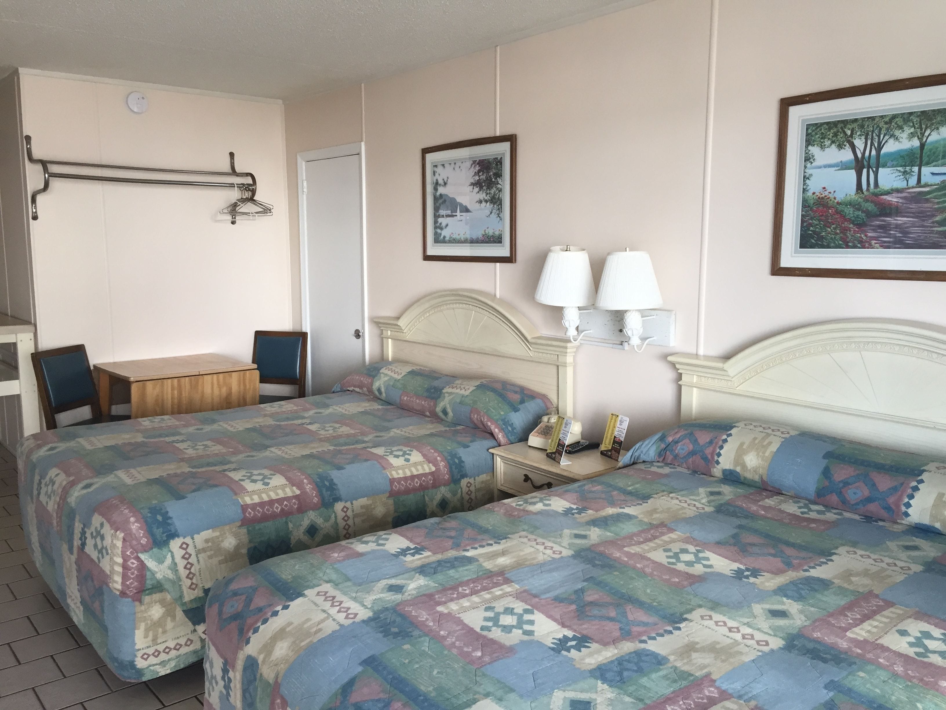 A hotel room with two beds, a desk, and a chair. There are two wall-mounted lamps and a framed picture above the bed.