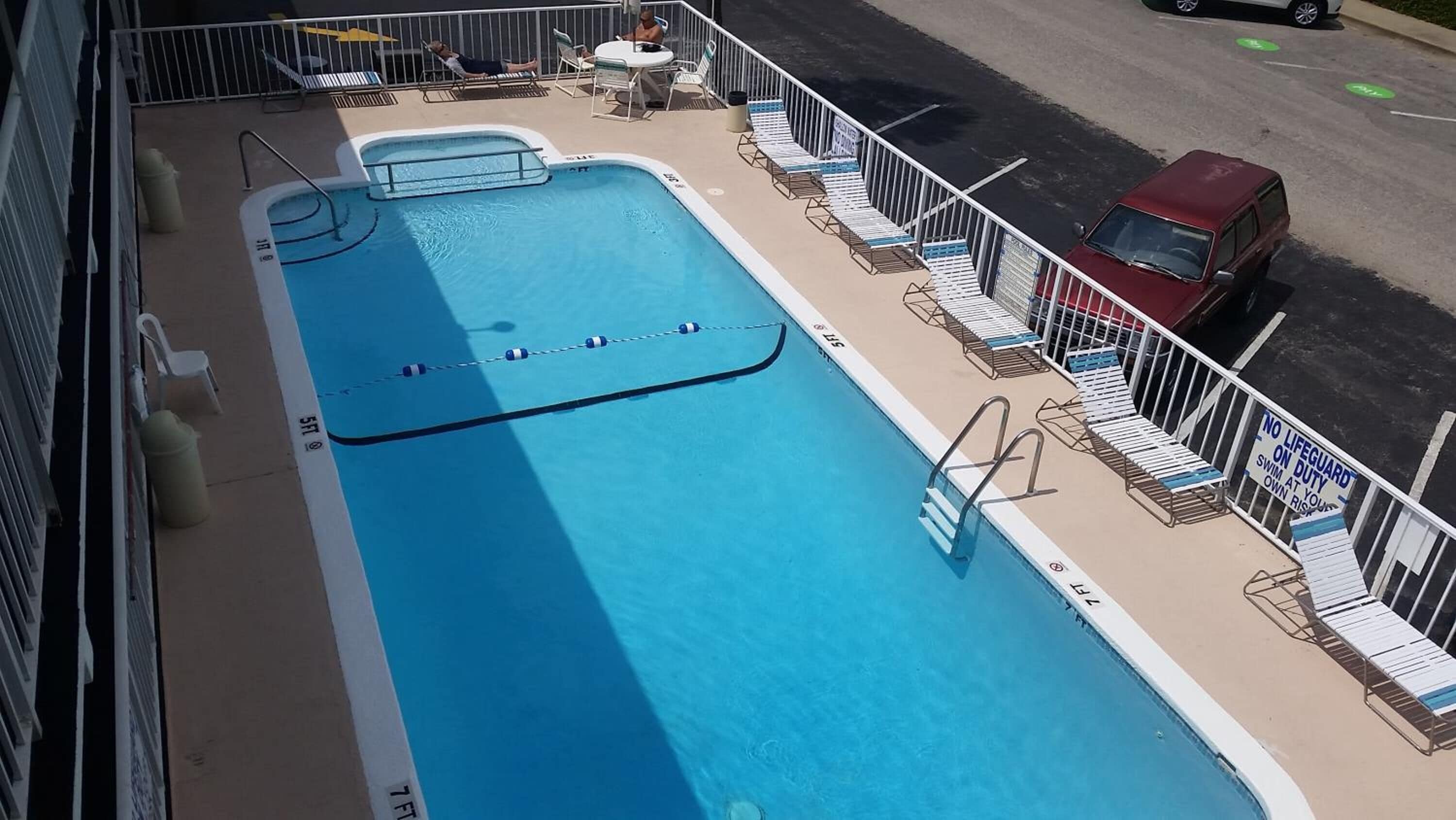 Un espace piscine avec des chaises longues, un panneau indiquant la présence d’un sauveteur et un véhicule rouge garé à proximité.