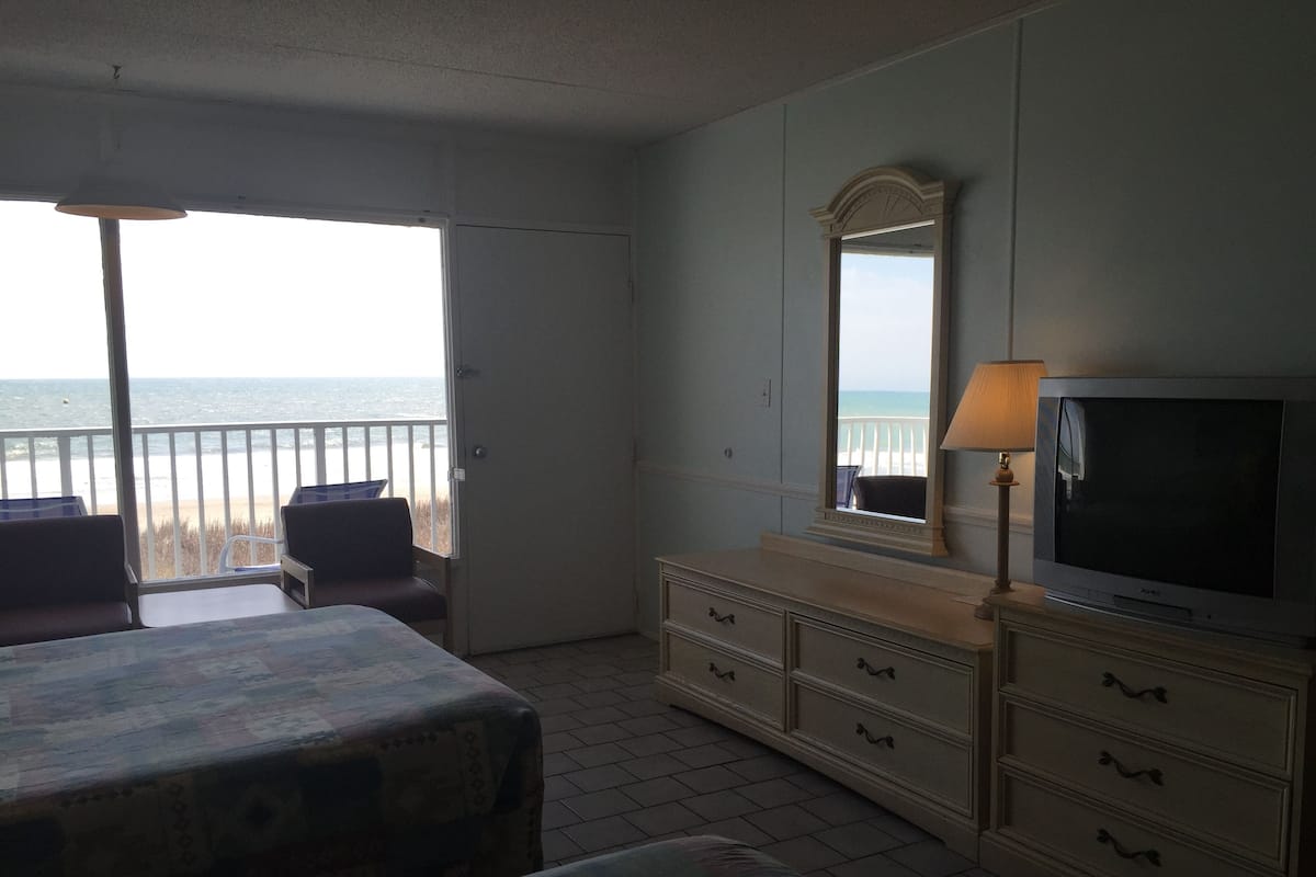 Quarto de hotel com cama, penteadeira, televisão e varanda com vista para o oceano.