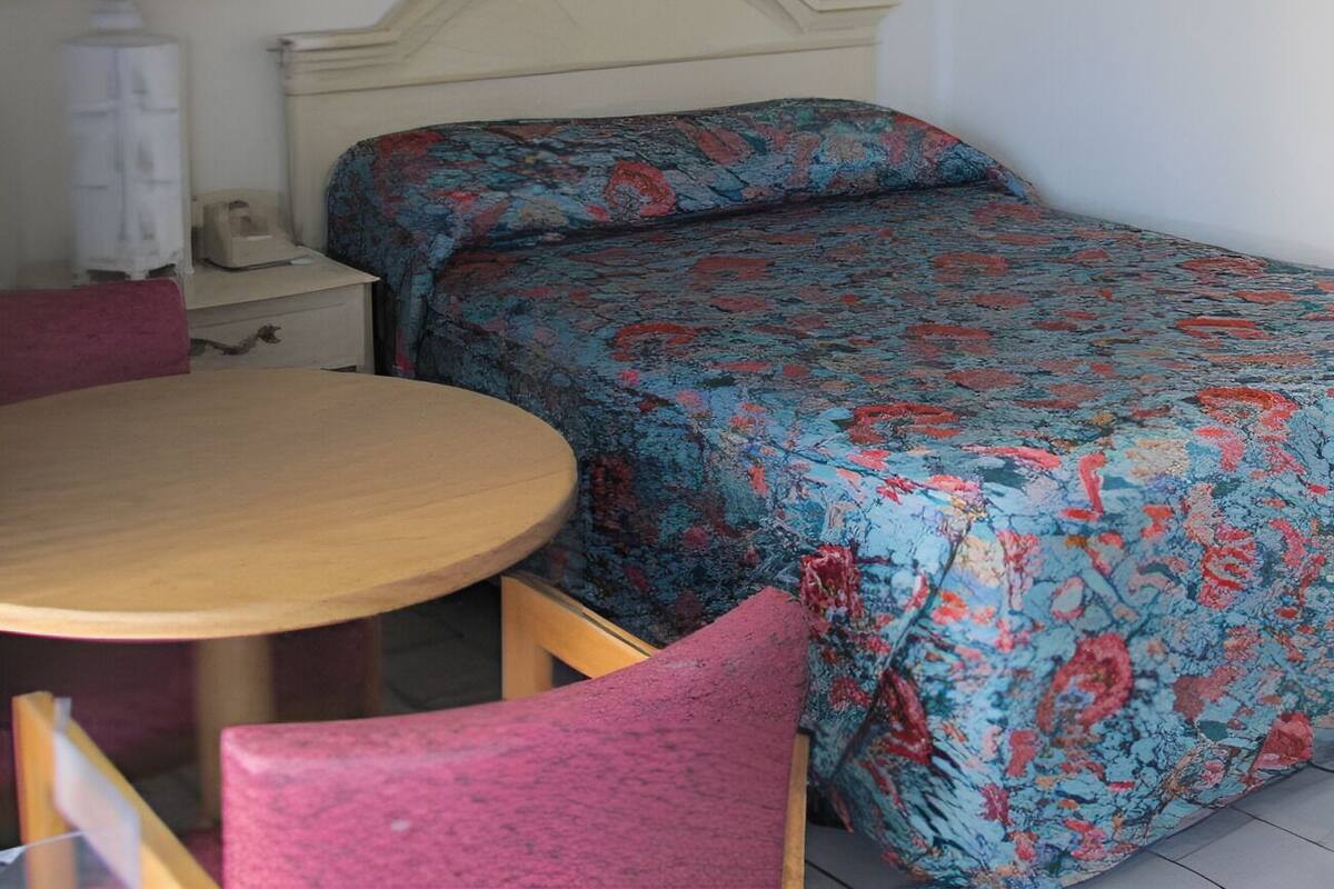 Um quarto pequeno e simples com uma cama, uma mesa redonda e uma cadeira.
