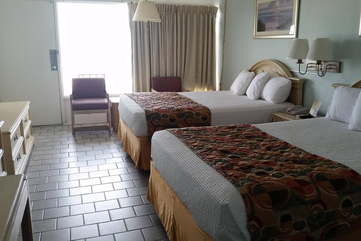 Quarto de hotel com duas camas, uma cadeira, uma escrivaninha e uma janela com cortinas.