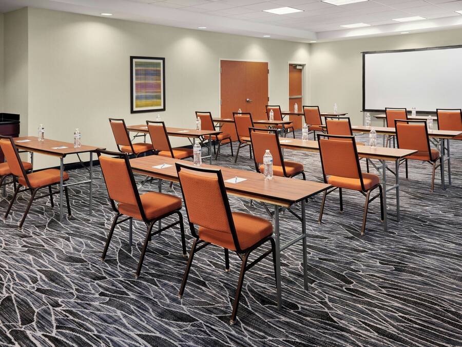 A conference room with multiple tables and chairs, equipped with water bottles, a projector screen, and a carpeted floor.