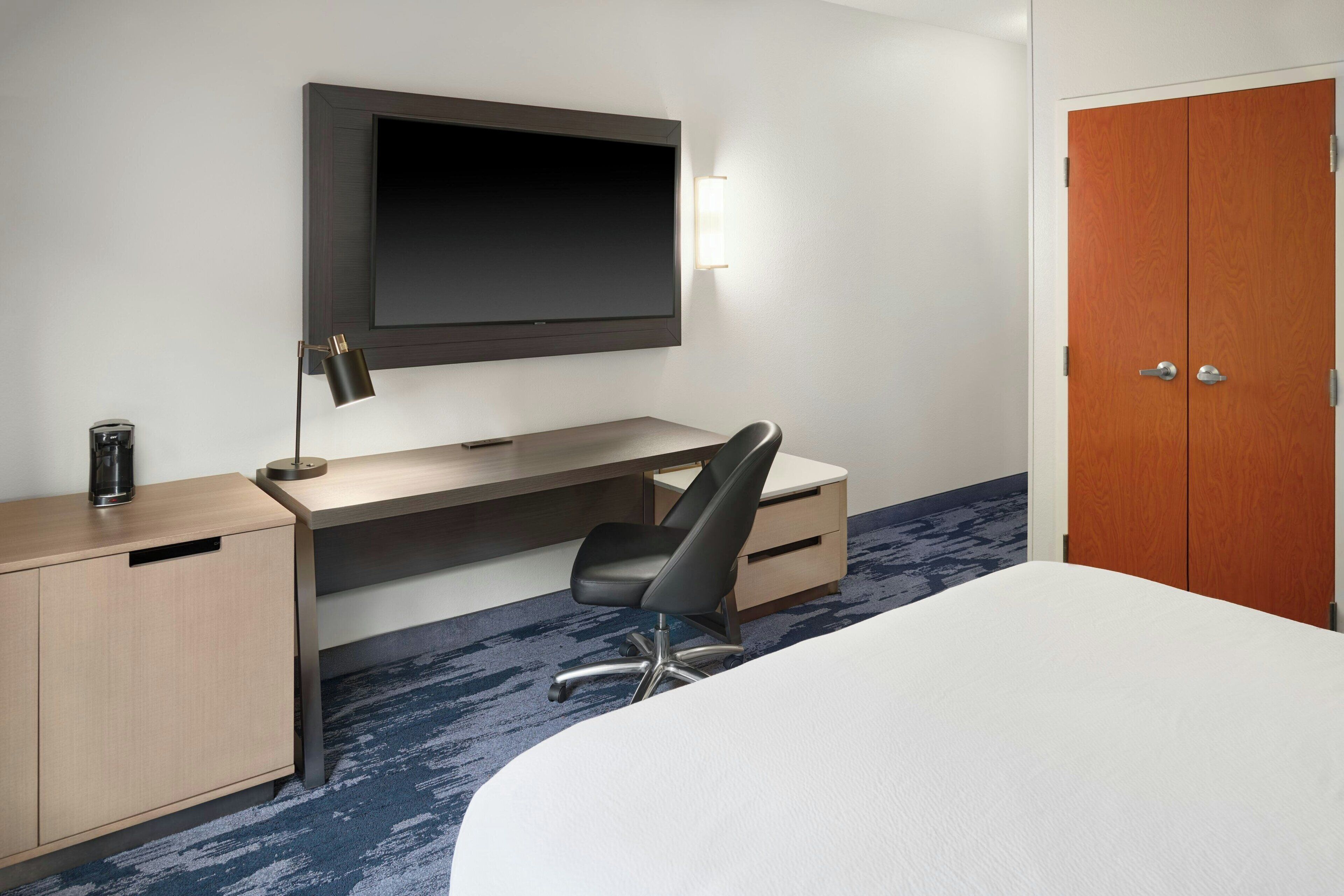 A hotel room with a bed, a desk with a chair, a television mounted on the wall, and a wardrobe.