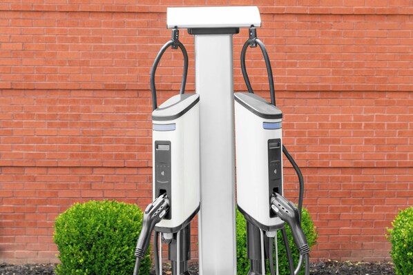 Duas estações de carregamento para veículos elétricos fixadas a um poste branco, com uma parede de tijolos ao fundo.