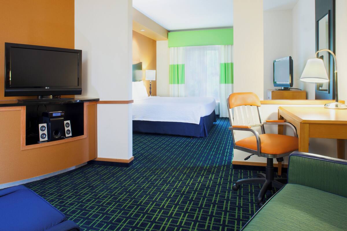 A hotel room with a bed, a desk, a chair, a television, and a small table with a lamp.