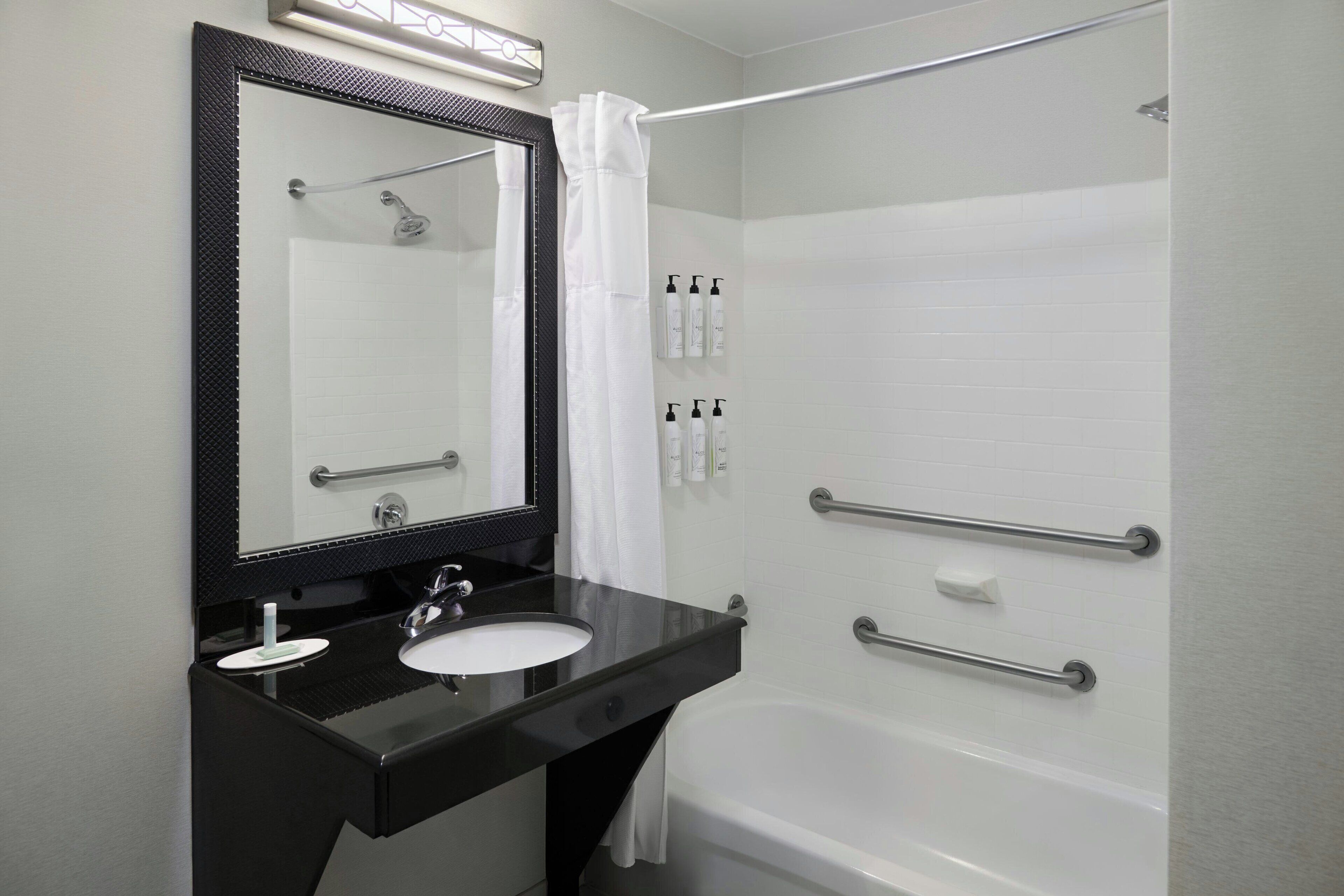 A bathroom with a black vanity, white sink, and a large mirror. There are handrails on the shower and a white bathtub.