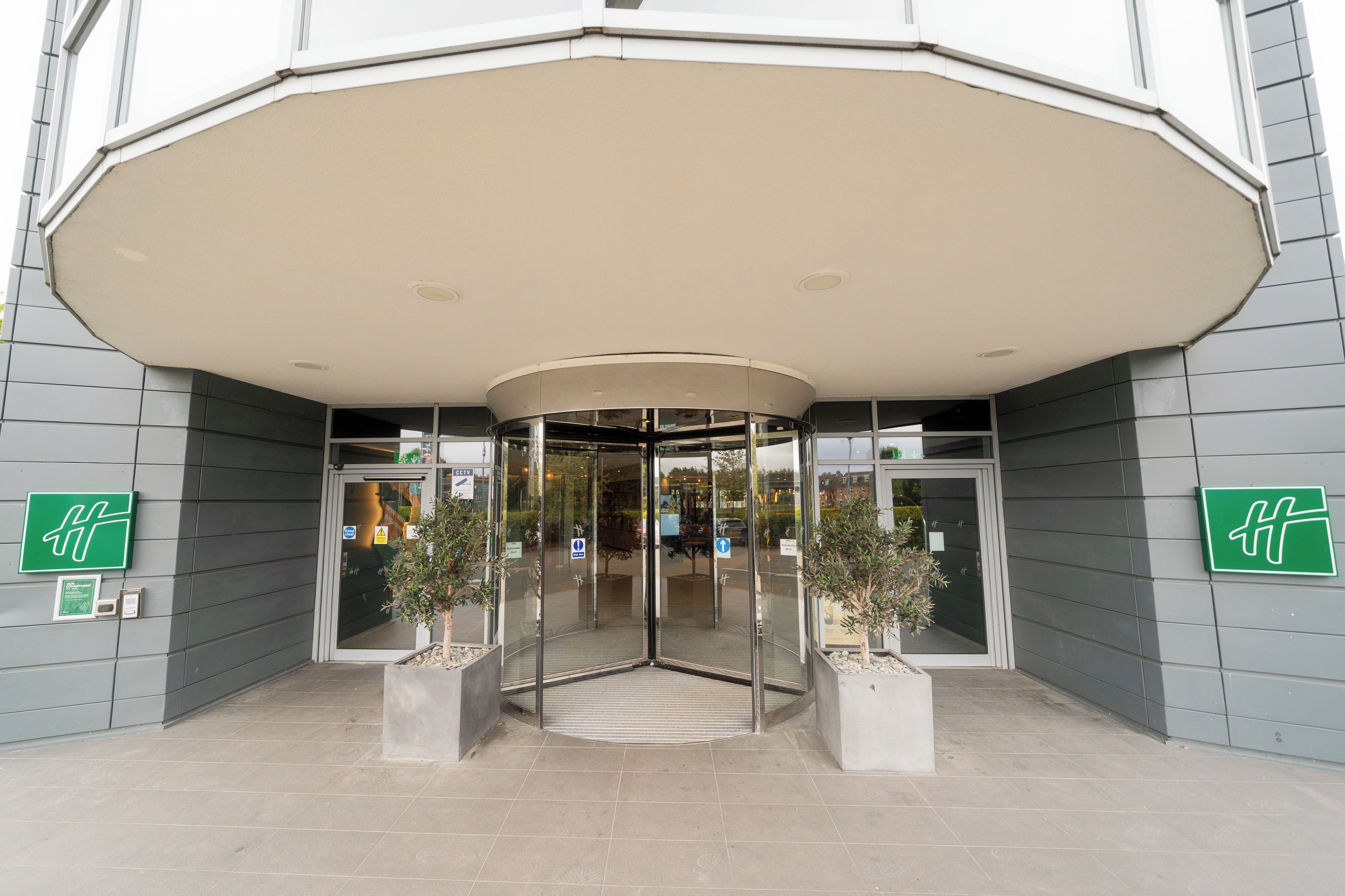 Entrada de hotel con puerta de cristal, zona de espera cubierta y plantas en macetas.