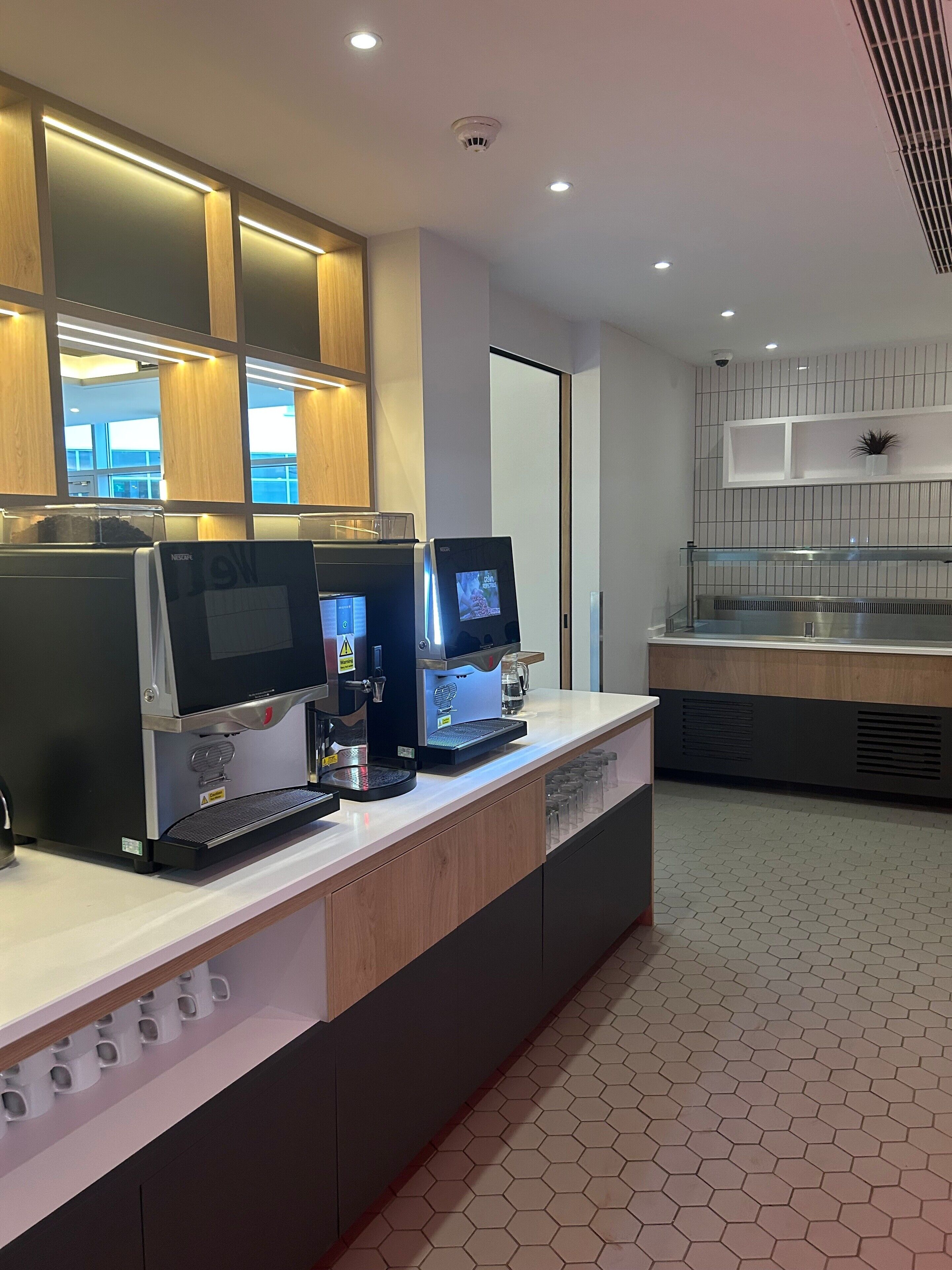 A modern kitchen area with two coffee machines, a counter with a glass display, and a hexagonal tiled floor.
