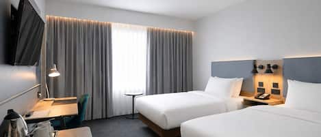 A hotel room with two beds, a desk with a chair, a TV mounted on the wall, and a bedside table with lamps.