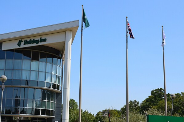 A Holiday Inn building with a modern glass facade, flagpoles with flags, and a clear blue sky.