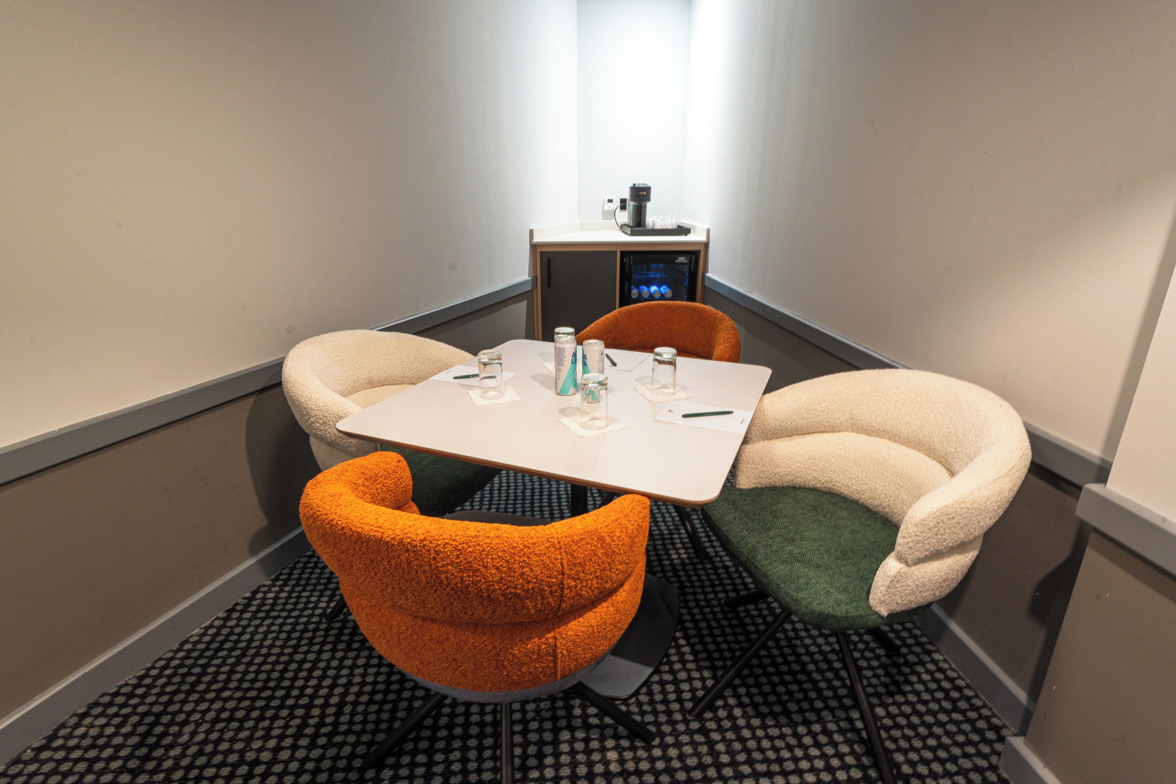 A small meeting room with a white table, two chairs, a coffee machine, and a refrigerator.