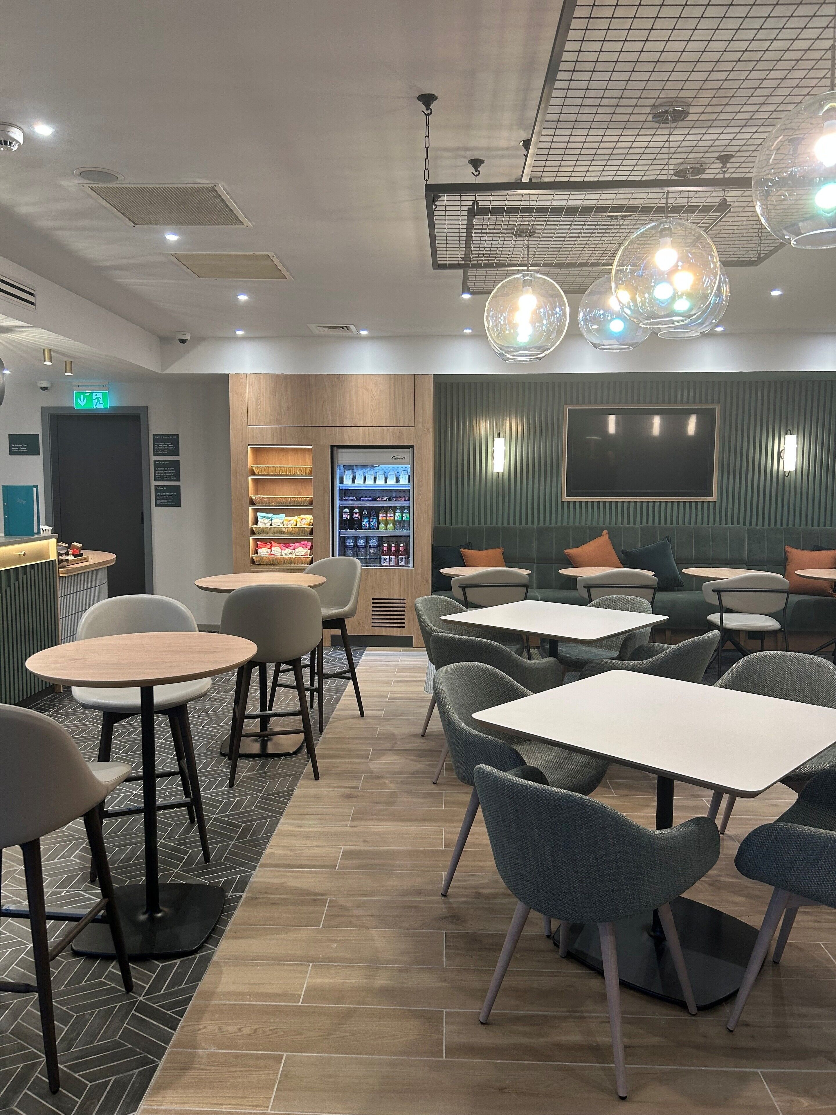 Interior de una cafetería moderna con mesas y sillas, un refrigerador con bebidas y un televisor en la pared.