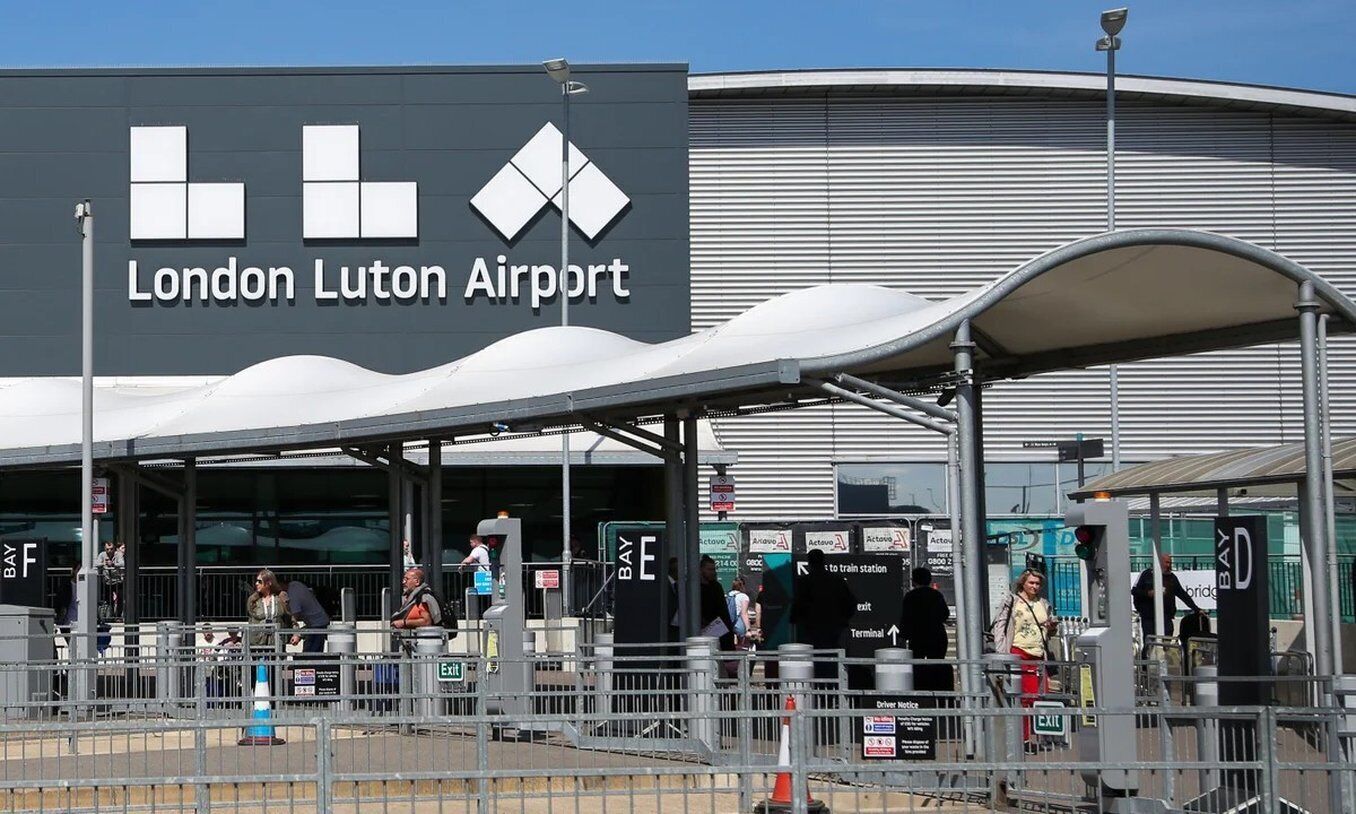 London Luton Airport with a large sign, a covered waiting area, and a metal fence.
