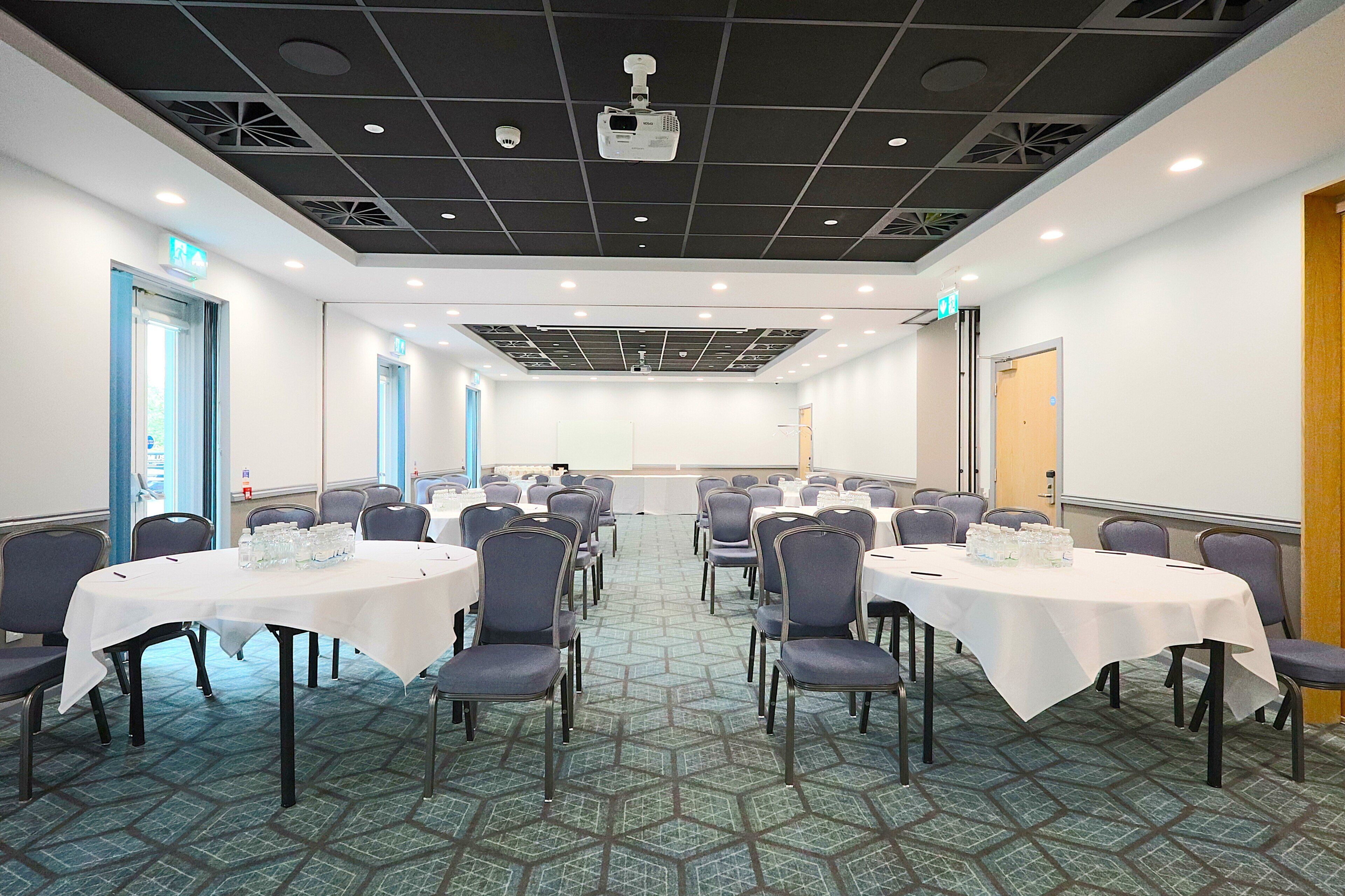 A conference room with multiple tables and chairs, equipped with a projector and a whiteboard.