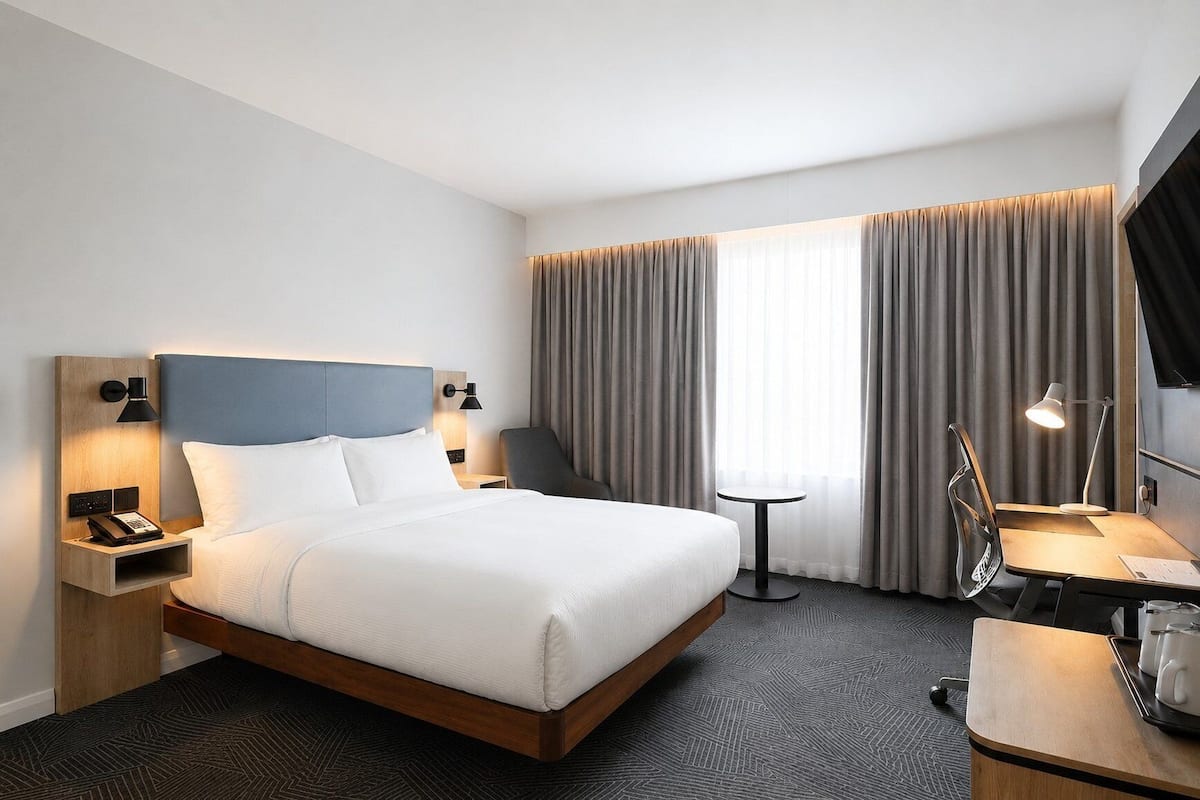 A hotel room with a bed, bedside table with a phone, a desk with a chair, a small table with a lamp, and a large window with curtains.
