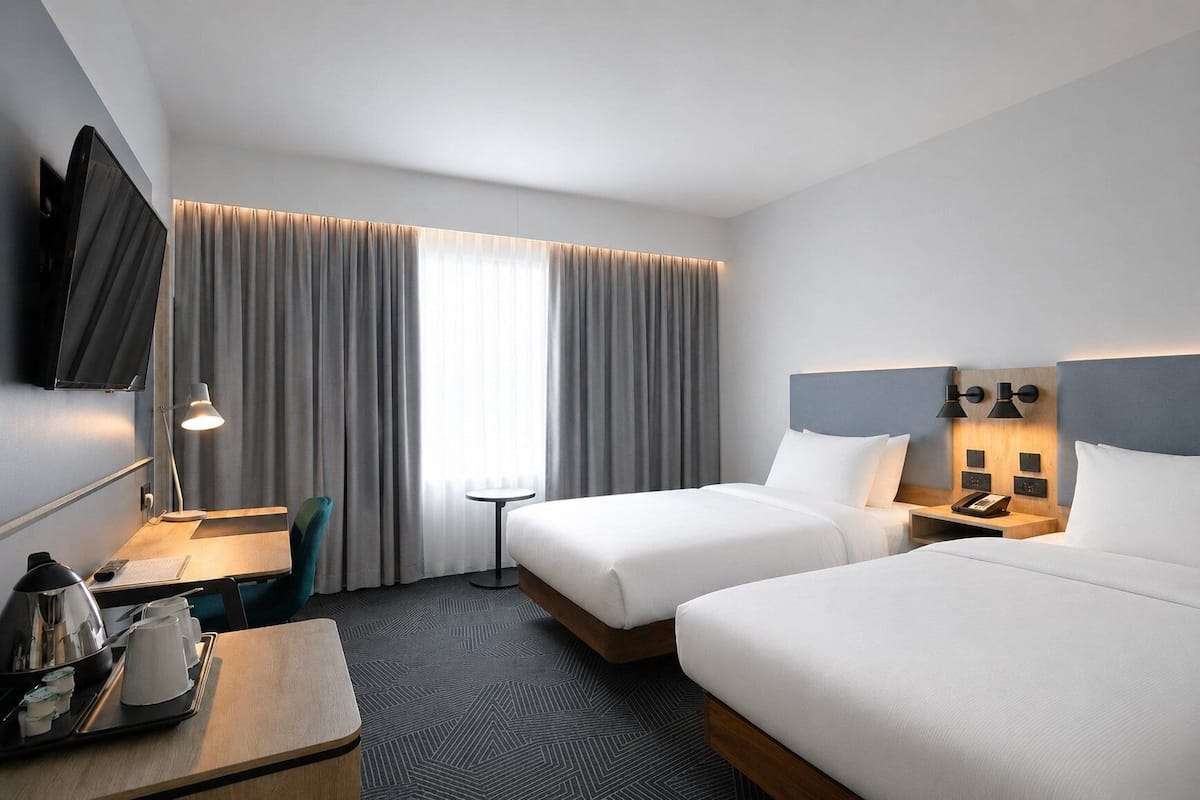 A hotel room with two beds, a desk with a chair, a TV mounted on the wall, and a bedside table with lamps.