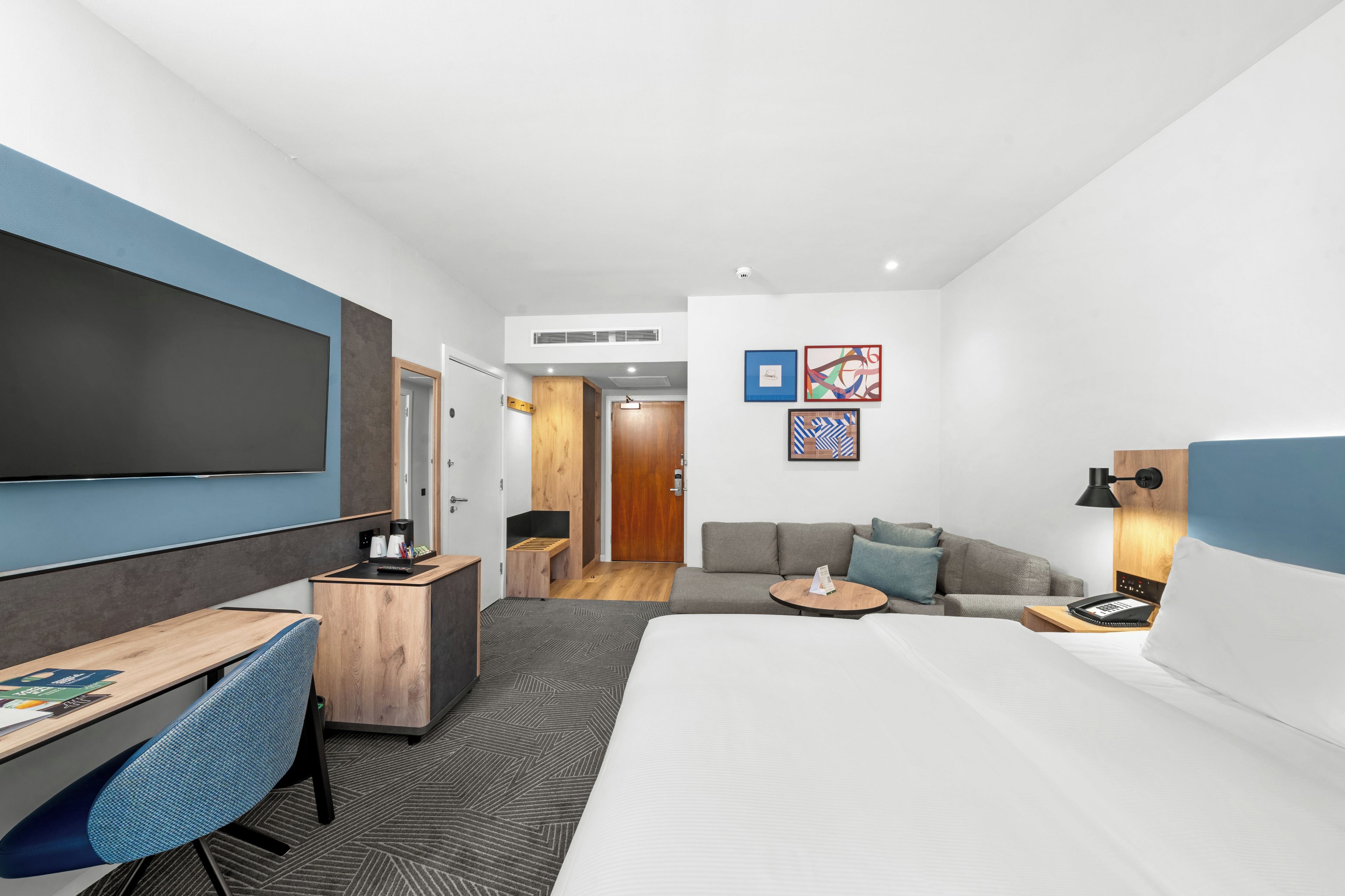 A modern hotel room with a large bed, a desk with a chair, a sofa, and a TV mounted on the wall.