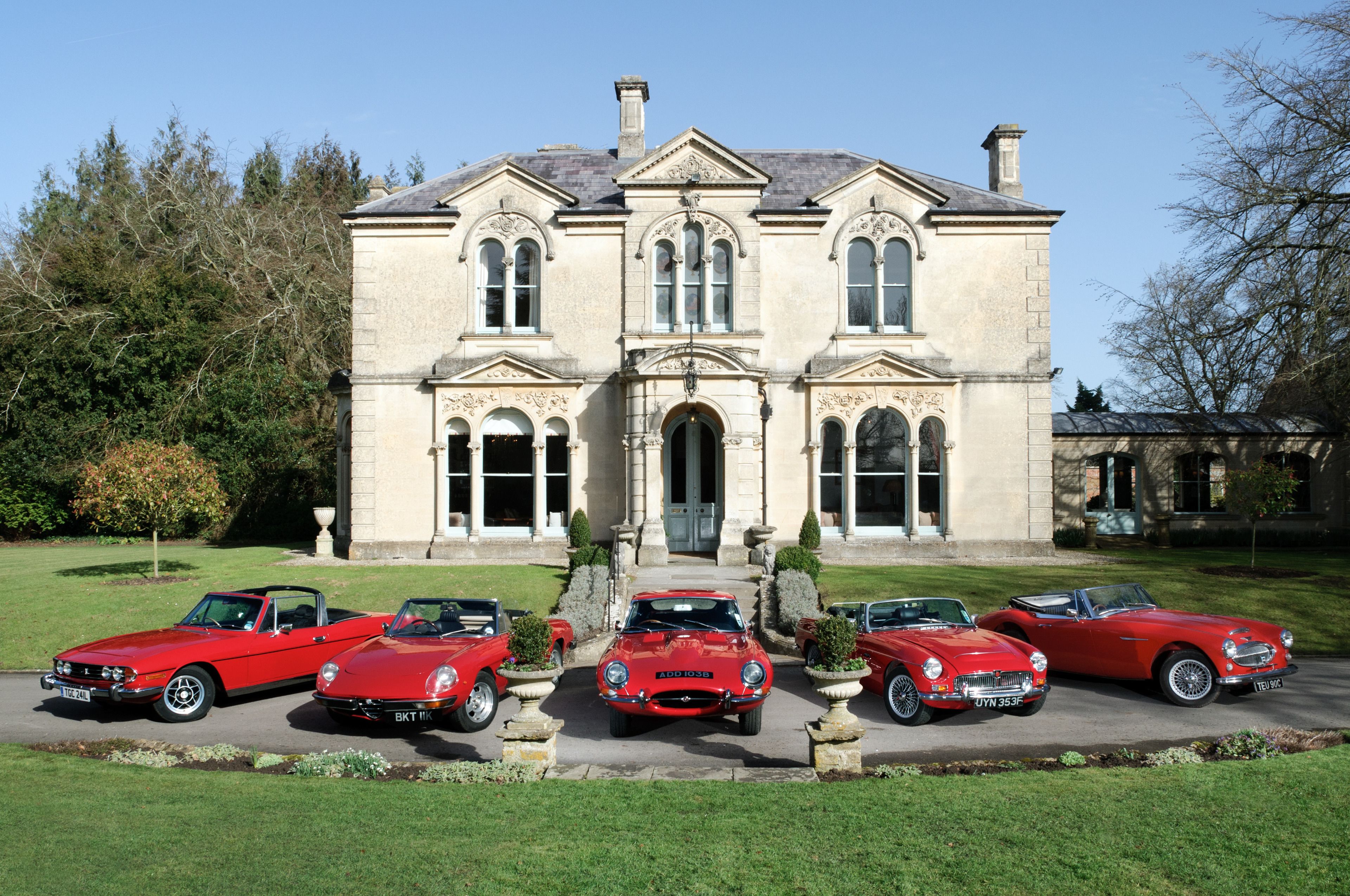 A large stately building with a symmetrical facade, a central entrance, and a driveway with four classic red cars parked in front.