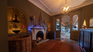Une pièce chaleureuse, éclairée d’une lumière douce, avec une cheminée, un tapis rouge à motifs et un sapin de Noël.