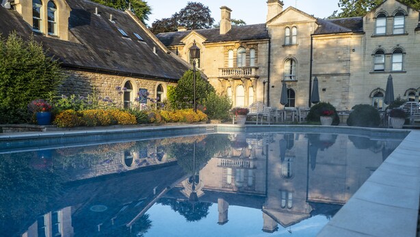 Une piscine dont l’eau reflète les bâtiments environnants, entourée de plantes en pot et de mobilier d’extérieur.