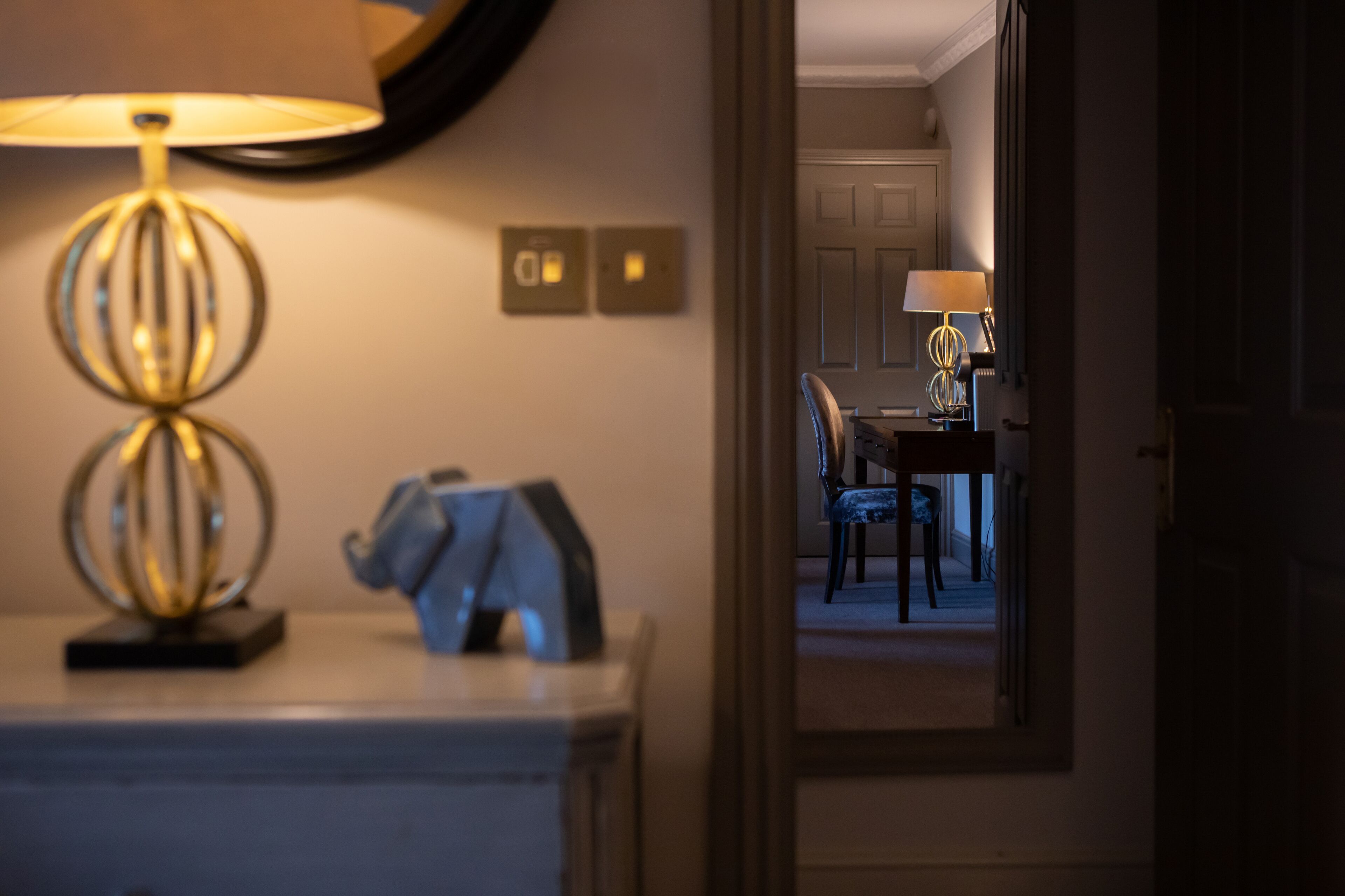 Habitación de hotel con lámpara de noche, mesita de noche y espejo que refleja un comedor con mesa y sillas.