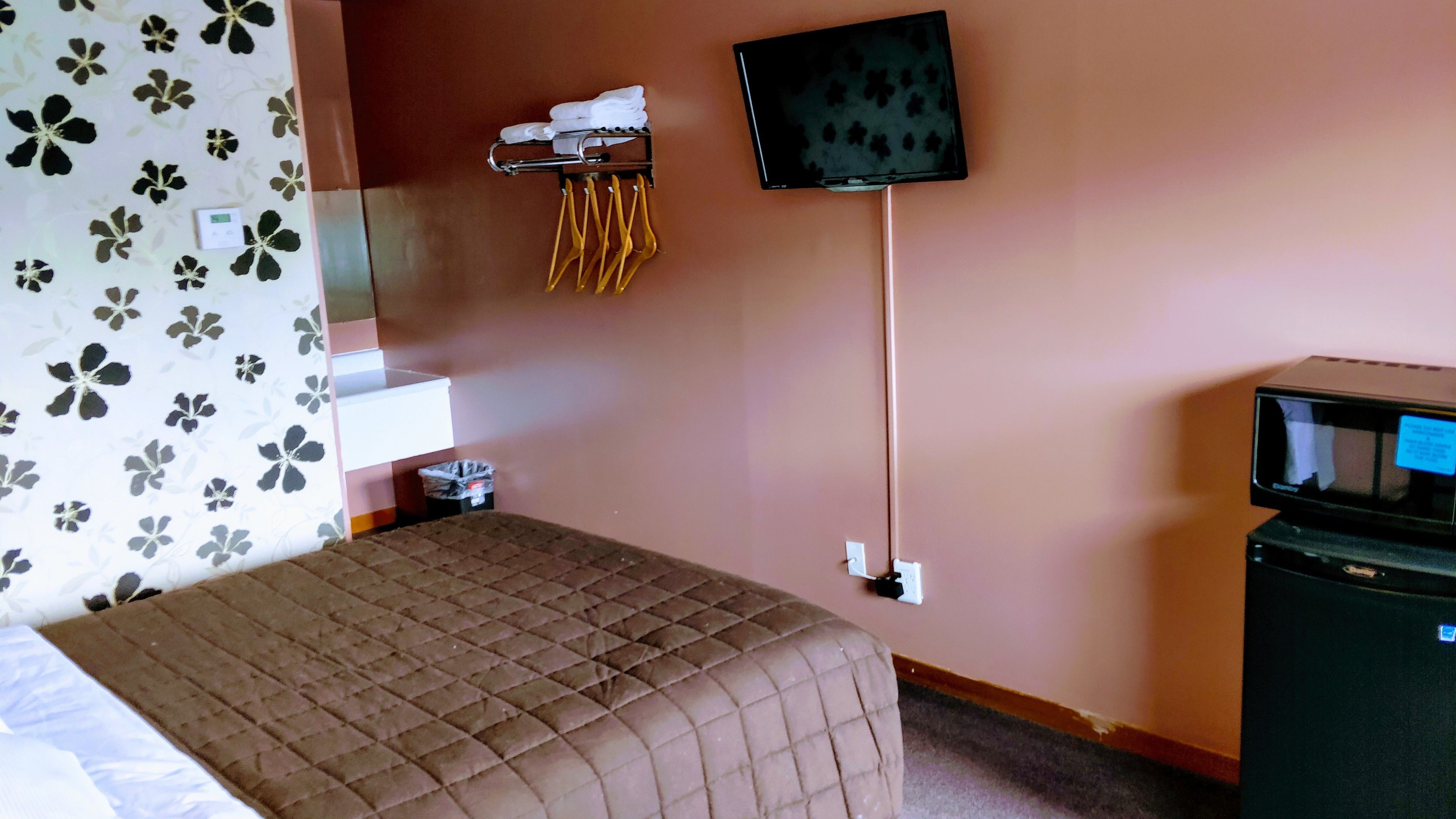A hotel room with a bed, a TV mounted on the wall, a small shelf with towels, and a trash bin.