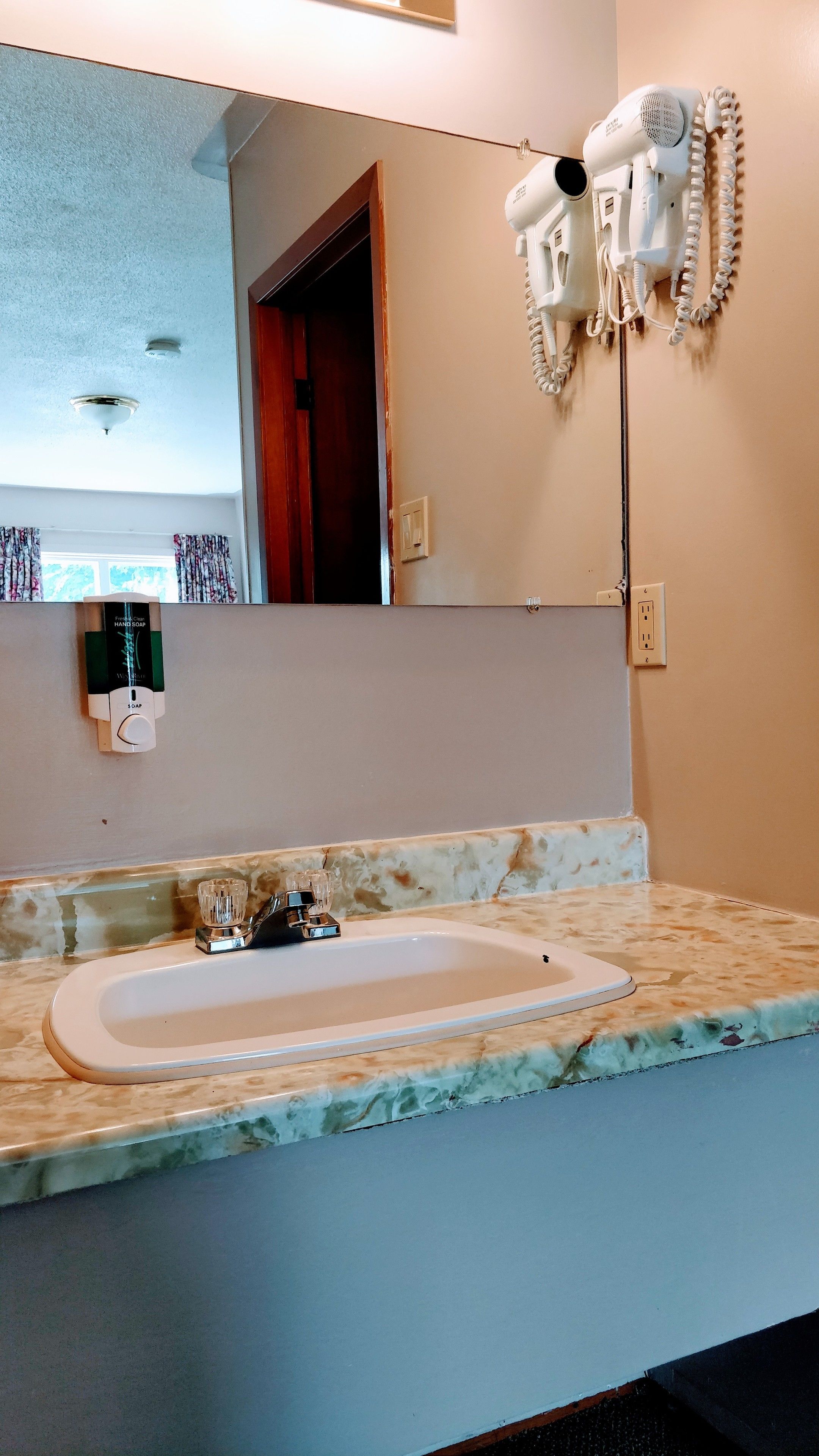 A bathroom with a marble countertop, a sink, a hand soap dispenser, a hairdryer mounted on the wall, and a mirror.