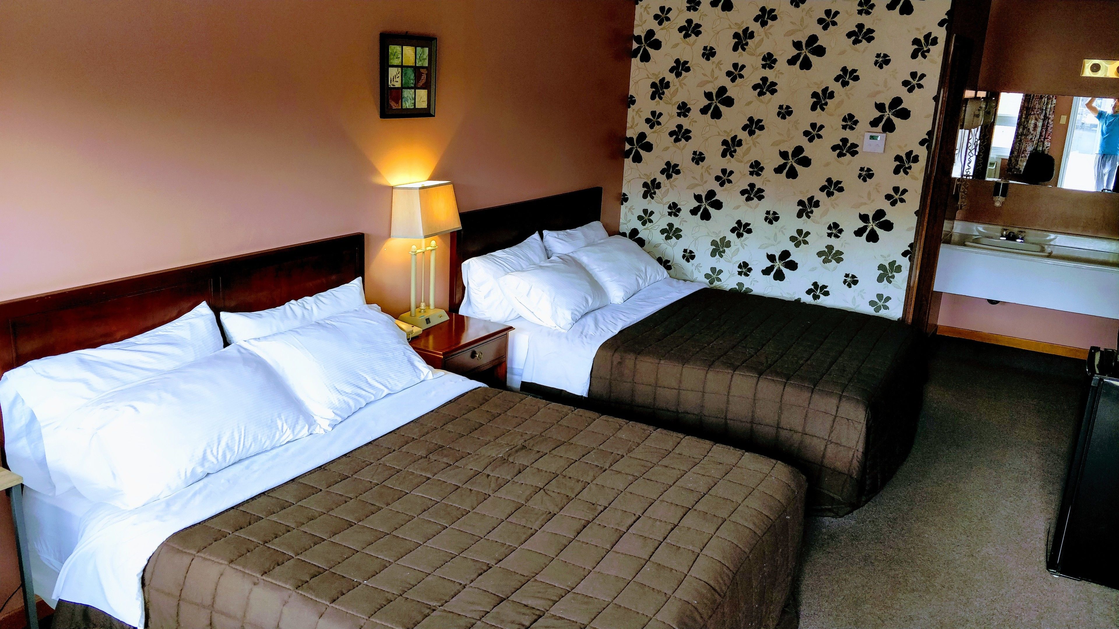 Camera d'albergo con due letti, un comodino con una lampada, uno specchio e una parete con motivo floreale.