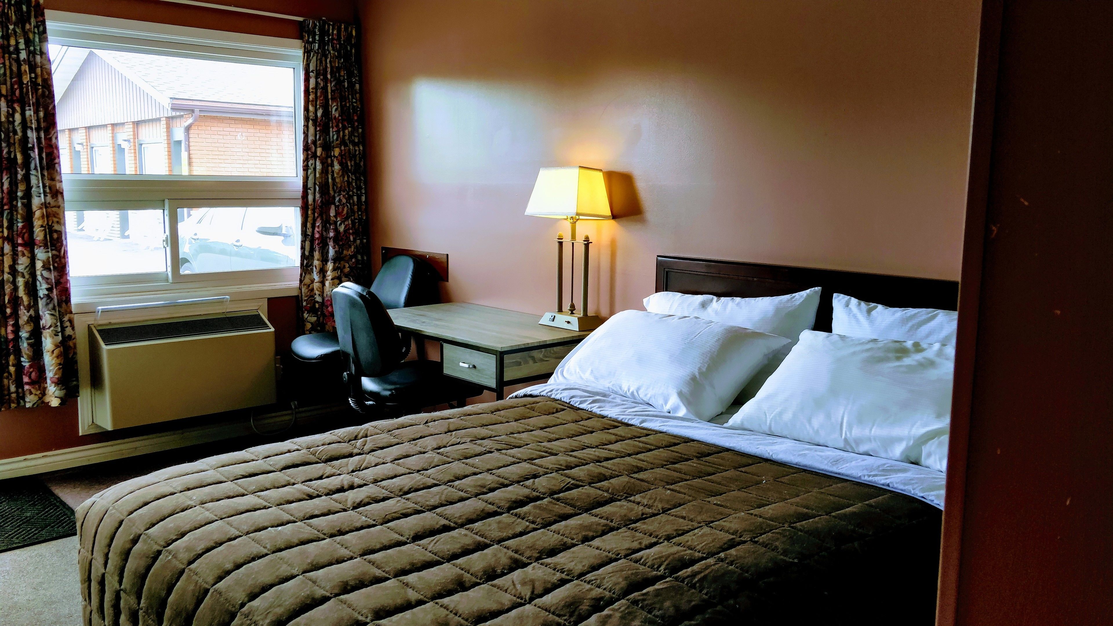 Ein Hotelzimmer mit Bett, Schreibtisch, Stuhl, Lampe und einem Fenster mit Vorhängen.